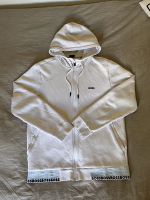 Vit hoodie med dragkedja från BOSS - Snygg vit hoodie från BOSS med dragkedja framtill och justerbar huva. Diskret BOSS-logga på bröstet och upprepade BOSS-tryck längs nederkanten. Mjuk bomullskänsla och två sidofickor. Perfekt för en clean och stilren look. Obs! Det finns en liten fläck i mitten av tröjan som endast syns på nära håll, inget man lägger märket till.
