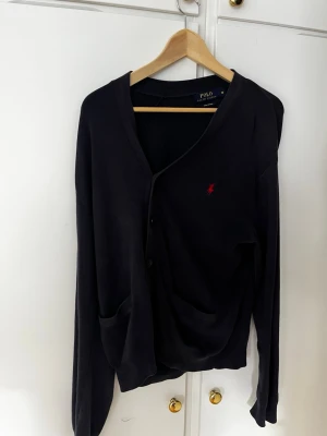 Svart cardigan från Polo Ralph Lauren - Svart cardigan med knäppning framtill från Polo Ralph Lauren. Klassisk design med två fickor och röd broderad logga på bröstet. Långärmad och i mjukt material, perfekt för lager-på-lager-stil.