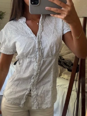 Vit prickig blus med volang - Söt vit blus med små svarta prickar och volangkant längs knäppningen. Blusen har korta ärmar och markerad midja. Perfekt till jeans eller kjol för en fräsch somrig look. Godkänner prisförslag vid snabb affär!💕