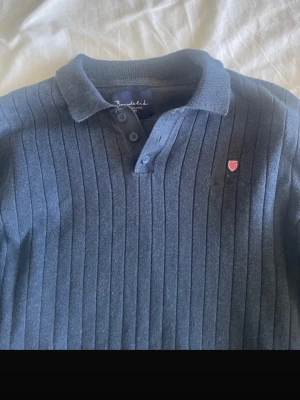 Mörkblå ribbad pikétröja Bondelid - Mörkblå långärmad pikétröja från Bondelid med ribbat mönster och liten röd logotyp på bröstet. Tröjan har klassisk krage och knäppning med tre knappar. Perfekt för en stilren och avslappnad look.