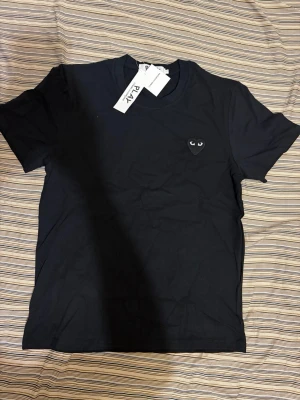 Cdg t shirt - Säljer en svart t-shirt från Comme des Garçons Play med det ikoniska hjärteögon-logotypen broderad på bröstet. T-shirten har rund hals, korta ärmar och är gjord i mjuk bomull. Perfekt för dig som gillar stilrena och trendiga plagg.