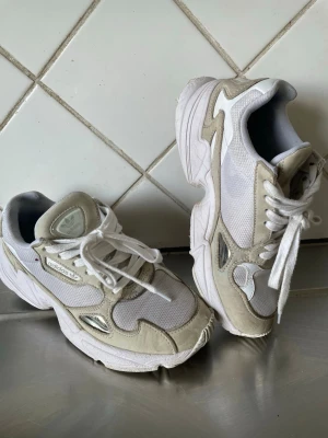Adidas Falcon vita sneakers - Chunky vita Adidas Falcon sneakers med mesh och mockadetaljer samt inslag av silver. Skorna har en bred sula och klassisk låg siluett. Perfekta för dig som gillar retrostil och streetwear-vibe. Snörning framtill och bekväm passform.