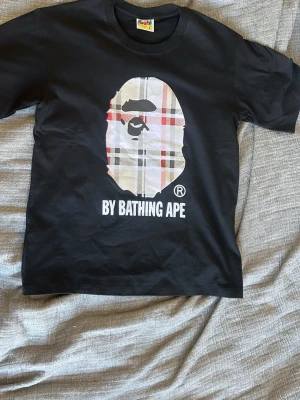 Svart BAPE t-shirt med rutig logga - Säljer en svart t-shirt från BAPE med det ikoniska apansiktet i rutig design på bröstet och texten 'BY BATHING APE' under. Klassisk passform och rund hals. Perfekt för dig som gillar streetwear och vill sticka ut. Använd endast 1 gång! Skriv vid intresse🙂