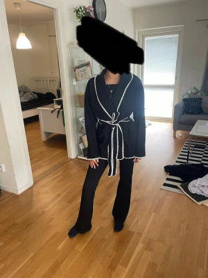 Svart cardigan med vit kant och knytskärp - Stilren svart cardiganjacka med markerade vita kanter och knytskärp i midjan. Jackan har en avslappnad passform, sjalkrage och långa ärmar. Perfekt för lager-på-lager och enkel att styla till olika outfits.