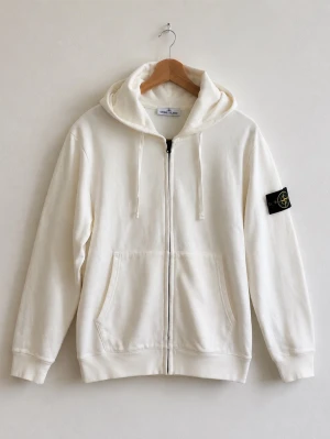 Vit hoodie från Stone Island - Snygg vit hoodie från Stone Island med dragkedja framtill, huva med dragsko och kängurufickor. Klassisk svart patch med logga på vänster ärm. Tillverkad i mjuk bomull för en skön känsla.