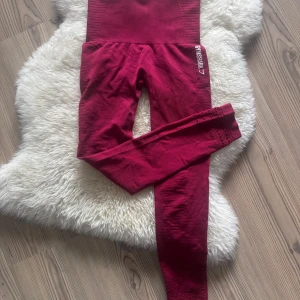 Röda seamless leggings från Gymshark - Snygga röda seamless leggings från Gymshark med hög midja och ribbade detaljer längs sidorna och midjan. Tight passform och diskret hålmönster vid bensluten. Perfekta för träning eller chill dagar.