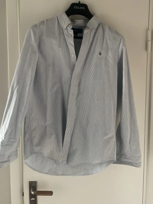 Randig skjorta från Ralph Lauren - Klassisk blå- och vitrandig skjorta från Ralph Lauren med knappar framtill och lång ärm. Skjortan har en liten broderad logga på bröstet och är tillverkad i bomull. Perfekt för en stilren look.