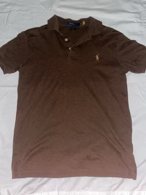 Brun pikétröja från Polo Ralph Lauren - Brun pikétröja från Polo Ralph Lauren med klassisk krage och knappslå framtill. Tröjan har korta ärmar och den ikoniska broderade loggan på bröstet. Perfekt för en avslappnad men ändå stilren look.