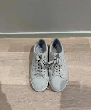 Vita Tommy Hilfiger sneakers - Snygga vita sneakers från Tommy Hilfiger med klassisk låg profil och diskret logga på sidan. Skorna har vita snören och ovandel i slätt material, perfekt för en clean och stilren look. Passar till det mesta och ger en fräsch vibe till outfiten.