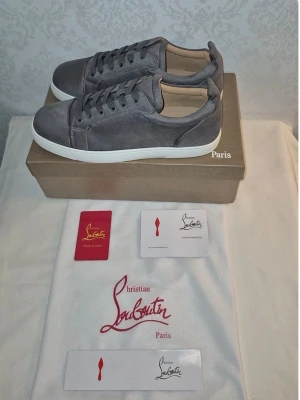 Gråa Christian Louboutin sneakers - Snygga gråa sneakers från Christian Louboutin i mocka med klassisk röd sula och vita kanter. Lågt skaft, rund tå och tonade skosnören. Perfekta för dig som vill ha en stilren och exklusiv look med en ikonisk touch. Helt nya. Passar perfekt till sommaren!Först till kvarn!