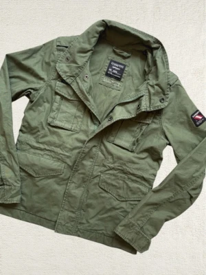 Grön utilityjacka från Superdry - Snygg militärgrön utilityjacka från Superdry med flera fickor framtill och patch på ena ärmen. Jackan har klassisk krage, dragkedja och knappar samt lång ärm. Perfekt för dig som gillar streetstyle och funktionella detaljer.