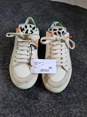 Beige sneakers från Axel Arigato - Snygga beige sneakers från Axel Arigato med coola detaljer som leopardmönstrad plös, grönt parti vid hälen och orange detaljer på skosnörena. Tillverkade i läder med mockadetaljer vid tån. Perfekta för dig som vill sticka ut med din stil.