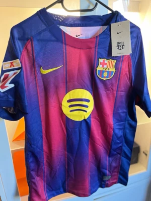 FC Barcelona matchtröja Nike S - Säljer en officiell FC Barcelona matchtröja från Nike med Dri-FIT material. Tröjan har blå och vinröda vertikala ränder, gula detaljer och klubbmärke på bröstet. Sponsorlogga från Spotify på framsidan och patch på ärmen. Perfekt för fotbollsfans!