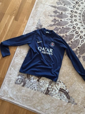 Mörkblå PSG träningströja Nike - Mörkblå långärmad träningströja från Nike med PSG-logga och Qatar Airways-tryck på bröstet. Tröjan har en sportig design med rund hals och är perfekt för fotbollsträning eller chill. Materialet känns lätt och andas bra.