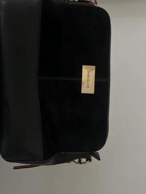 Svart axelväska River Island - Snygg svart axelväska från River Island med guldfärgad logga och dragkedja. Väskan har en mix av slätt och mockaliknande material samt en beige tofsdetalj i sidan. Perfekt storlek för det viktigaste och justerbar axelrem.