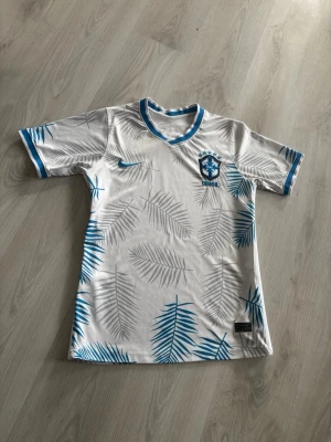 Vit, ljusblå Brasilien fotbollströja Nike S - Snygg vit och blå  fotbollströja från Nike med Brasilien-emblem på bröstet. Tröjan har blå detaljer vid krage och ärmslut samt ett mönster av palmblad i blått och grått över hela plagget. Perfekt för dig som älskar fotboll och vill sticka ut på planen. Sitter som S