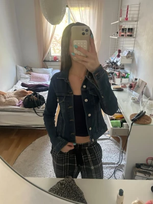 Mörkblå jeansjacka med silverknappar - Snygg mörkblå jeansjacka med klassisk krage och silverfärgade knappar framtill. Jackan har två bröstfickor med lock och knapp, samt figurnära passform som slutar vid midjan. Perfekt att slänga över en topp för en cool look.