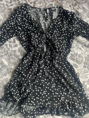 Svart prickig kortklänning/ byxdress med volang - Kort svart klänning med vita prickar och volangkant nertill. Klänningen har v-ringning med rynkdetalj framtill, markerad midja och trekvartsärm. Tillverkad i ett lätt och luftigt material som ger en skön känsla.