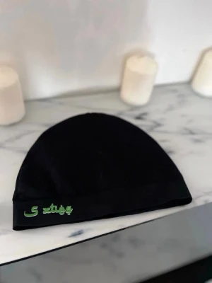 Svart mössa med grön brodyr - Svart mössa med broderad grön text framtill. Enkel och stilren design som passar till de flesta outfits. Mjuk och stretchig, perfekt för kyliga dagar.