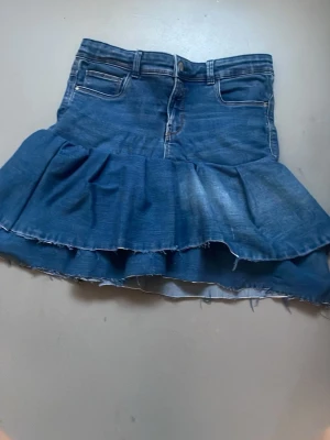 Jeans Volangkjol - Säljer en blå jeanskjol med volang. Kjolen har klassiska fickor fram och bak, knapp och dragkedja i midjan är den stretchig och jätteskön. Perfekt för dig som letar efter en snygg kjol till i sommar.