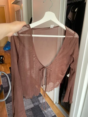 Brun mesh bolerotopp med knyt - Trendig brun bolerotopp i mesh med lång ärm och v-ringning. Toppen är croppad och har knyt framtill för en snygg detalj. Perfekt att styla över ett linne eller topp för en cool lager-på-lager-look.