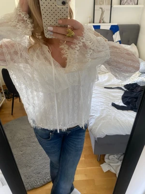 Vit spetsblus med volangdetaljer - Superfin vit blus i spets med romantiska volangdetaljer och knytband vid halsen. Blusen har långa, transparenta ärmar och ett luftigt fall. Perfekt för dig som gillar en feminin och trendig stil.
