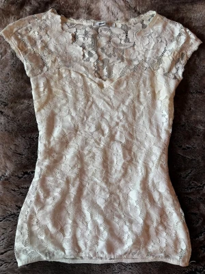 Vit spetsblus från Gina Tricot - Supersöt vit blus från Gina Tricot i spets med v-ringning och korta ärmar. Blusen har ett  blommigt spetsmönster och är perfekt att styla med jeans eller kjol. Lätt och luftig känsla, passar dig som gillar feminina detaljer.