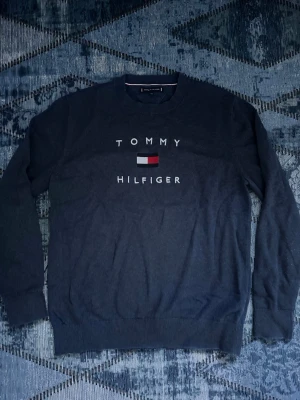Mörkblå Tommy Hilfiger tröja - Mörkblå långärmad tröja från Tommy Hilfiger med rund halsringning. Framsidan har det klassiska Tommy Hilfiger-logotyptrycket i vitt, rött och blått. Tröjan har ribbade muddar vid ärmslut och nederkant. Perfekt för en stilren och avslappnad look.