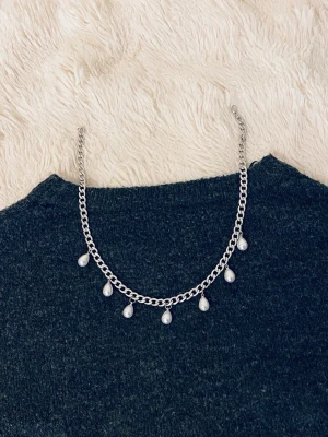 Silverfärgat halsband med pärldroppar - Snyggt halsband i silverfärgad metall med sju droppformade pärlor som hänger längs kedjan. Kedjan har en chunky design och pärlorna ger en elegant touch till looken. Perfekt för att lyfta vilken outfit som helst.
