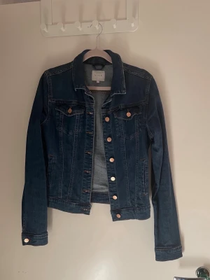 Mörkblå jeansjacka från Vila - Snygg mörkblå jeansjacka från Vila med klassisk krage, bröstfickor med knappar och kopparfärgade detaljer. Figurnära passform och långa ärmar. Perfekt att slänga över en hoodie eller t-shirt för en cool look. Lite liten i storlek är mer som stl S