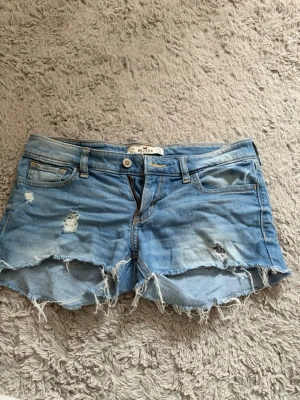 Blå slitna jeansshorts Hollister - Säljer ett par ljusblå jeansshorts från Hollister med slitna detaljer och fransig kant. Klassisk femficksmodell med låg midja och råa kanter för en avslappnad look. Perfekta för sommaren!