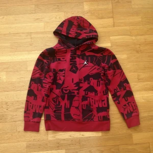 Röd Jordan hoodie med grafiskt tryck - Snygg röd hoodie från Jordan med svart grafiskt tryck över hela tröjan och den klassiska Jumpman-loggan på bröstet. Hoodien har huva och ribbade muddar vid ärmar och nederkant. Tillverkad i mjukt material som känns skönt mot huden.
