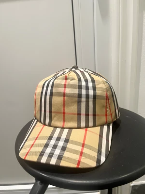 Burberry keps  - Snygg keps från Burberry med klassiskt rutigt mönster i beige, svart, vitt och röda detaljer. Märke med 'Burberry London England' baktill. Perfekt accessoar för dig som vill ha en lyxig och trendig look. Justerbar baktill för bästa passform.