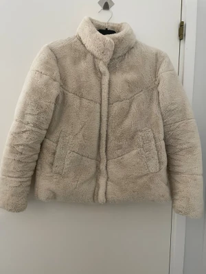 Beige teddyjacka från Zara XS - Supermjuk och fluffig teddyjacka från Zara i beige färg. Jackan har hög krage, två fickor framtill och stängs med dold dragkedja. Perfekt för kalla dagar när du vill vara både varm och stylish. Har en svag fläck som är knappt synbar samt en på kanten av ena armen. 