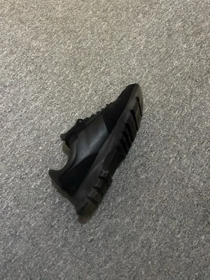 Svarta Prada sneakers med chunky sula - Snygga helsvarta Prada sneakers med chunky sula och ikonisk metallogo på sidan. Skorna har en mix av läder och textil, klassisk snörning och låg profil. Perfekta för dig som vill ha en stilren men edgy look.