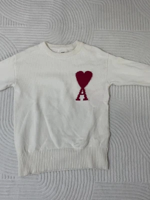 Vit stickad tröja från Ami Paris - Vit stickad tröja från Ami Paris med rund halsringning och ribbade muddar. På bröstet finns ett rött hjärta och bokstaven 'A' i samma färg, vilket är signaturdetaljen från märket. Perfekt för dig som gillar stilrena plagg med en twist. Storlek M men passar mest S