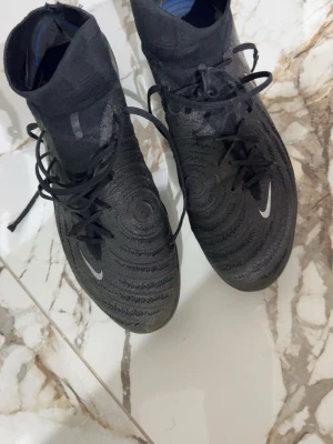 Svarta Nike Mercurial fotbollsskor - Säljer ett par svarta Nike Mercurial fotbollsskor med högt skaft och snörning. Skorna har en stickad ovandel med mönstrad textur för bättre bollkontroll och en vit Nike-logga på sidan och framtill. Perfekta för dig som vill ha stilrena och funktionella skor på planen.
