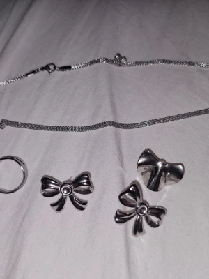 Smyckespaket med rosetter i silverfärg - Säljer ett smyckespaket med silverfärgade accessoarer: två rosettformade hängen, en enkel ring och en tunn kedja. Alla delar har en blank finish och rosetterna har en söt, ungdomlig design. Perfekt för dig som gillar detaljer i metall och vill mixa och matcha.