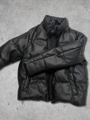 Zara jacka - Säljer en svart pufferjacka i fejkläder med hög krage och dragkedja framtill. Jackan har två sidofickor och en boxig, oversized passform som ger en cool streetwear-vibe. Perfekt för kalla dagar när du vill se stilren ut.