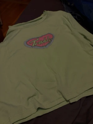 Ljusgrön croppad t-shirt Trouble - Säljer en ljusgrön croppad t-shirt med trycket 'Trouble' i rosa och vitt på bröstet. T-shirten har korta ärmar och är gjord i ribbad bomull som ger en skön och trendig känsla.