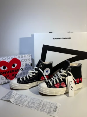 Converse x CDG Play svarta sneakers - Svarta höga sneakers från Converse i samarbete med Comme des Garçons Play. Skorna har vita sulor, vita snören och flera röda hjärtan med ögon på sidan. Klassisk Converse-logga på insidan och canvasmaterial för en cool streetstil.
