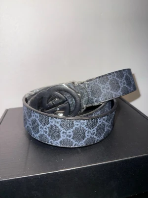 Svart GG bälte  - Snyggt svart bälte från GG med klassiskt GG-monogram i grått. Bältet har en rundad spänne och är gjort i skinn med tryckt mönster. Perfekt accessoar för att lyfta din outfit. 110CM verktyg för att justera storlek följer med 