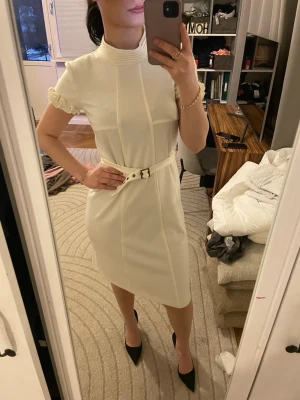 Vit kortärmad klänning med bälte - Stilren vit klänning med hög krage, korta ärmar med volangkant och markerad midja med matchande bälte. Klänningen har en rak passform och slutar vid knät. Perfekt för dig som gillar en klassisk och elegant look. Liknar Maje klänning😊