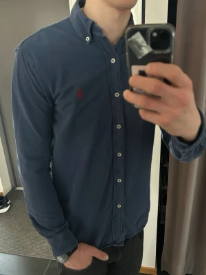 Mörkblå skjorta från Ralph Lauren - Klassisk mörkblå skjorta från Ralph Lauren med knappkrage och den ikoniska röda logotypen broderad på bröstet. Skjortan har långa ärmar, vita knappar och är tillverkad i mjuk bomull. Perfekt för en clean och stilren look.