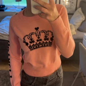 Rosa stickad tröja  - Cool rosa stickad tröja med svart kronmotiv på bröstet och text längs ena ärmen. Tröjan har rund halsringning och ribbade muddar. Perfekt för dig som vill sticka ut med en unik design.