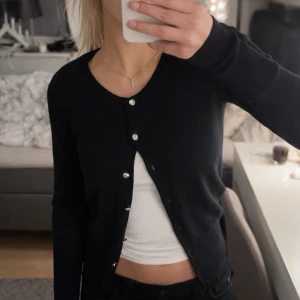 Svart kofta med glittriga knappar - Snygg svart kofta med långa ärmar och glittriga, dekorativa knappar framtill. Koftan har en smal passform och är tillverkad i ett ribbstickat material som ger en stilren look. Perfekt att bära över ett linne eller t-shirt.