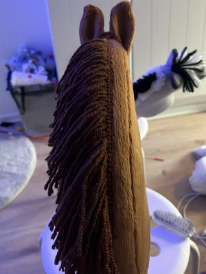 Brun käpphäst med garnman - Säljer en handgjord käpphäst i mjukt brunt tyg med mörkbrun man av garn och svarta broderade detaljer för ögon och näsborrar. Hästen har två upprättstående öron och en vit detalj vid ögat. Perfekt för lek och fantasi.