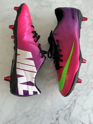Nike Mercurial rosa fotbollsskor - Snygga Nike Mercurial fotbollsskor i en grym rosa och lila färg med stor vit logga på sidan och en neon-grön swoosh. Skorna har svarta snören och metall-dobbar för bästa grepp på planen. Ovandelen är i syntetmaterial som ger bra bollkontroll och lätt känsla.