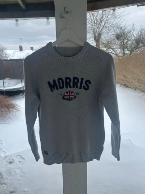 Morris Stockholm – Stickad tröja i nyskick (Grå) – Storlek M - Klassisk ljusgrå stickad tröja från Morris Stockholm i absolut nyskick. Inga anmärkningar, fläckar eller noppor. • Märke: Morris (Tailoring/Orange) • Storlek: Medium (M) • Färg: Gråmelange • Detaljer: Broderad logo med Union Jack på bröstet samt logo-patch nertill. Ribbstickade muddar.   