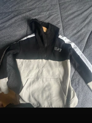 EA7 hoodie svart/grå med vit rand - Hoodie från EA7 i svart och ljusgrått med vit rand längs ärmarna. Halvlång dragkedja framtill, stor huva och magficka. EA7-logga på bröstet. Tillverkad i mjuk bomullsblandning för skön känsla och avslappnad stil.